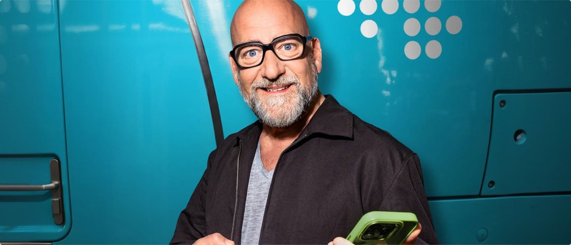 Mann mit Brille und grauem Bart hält ein Smartphone vor einem türkisfarbenen NAH.SH-Zug