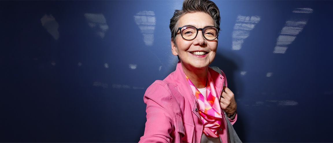 Kurzhaarige Frau mit Brille und pinker Jacke vor dunkelblauem Hintergrund