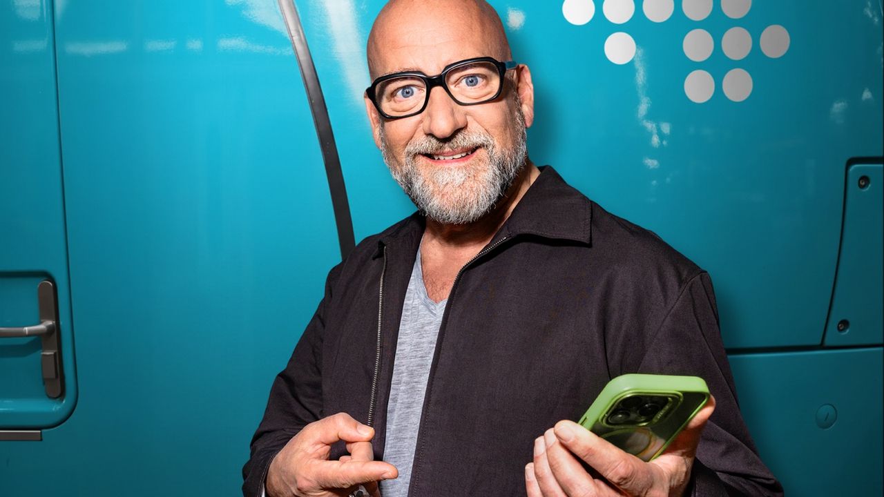 Mann mit Brille und grauem Bart hält ein Smartphone vor einem türkisfarbenen NAH.SH-Zug