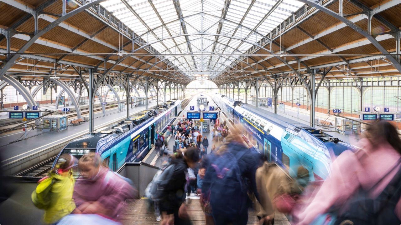 Reisende auf einem überdachten Bahnsteig mit einfahrendem NAH.SH-Zug