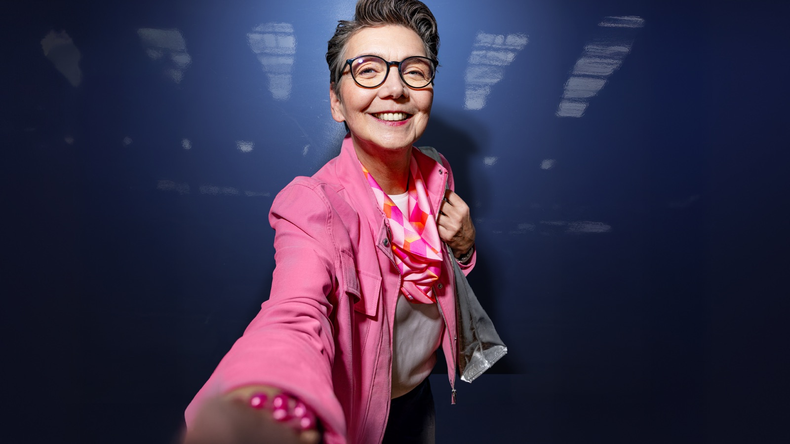 Kurzhaarige Frau mit Brille und pinker Jacke vor dunkelblauem Hintergrund